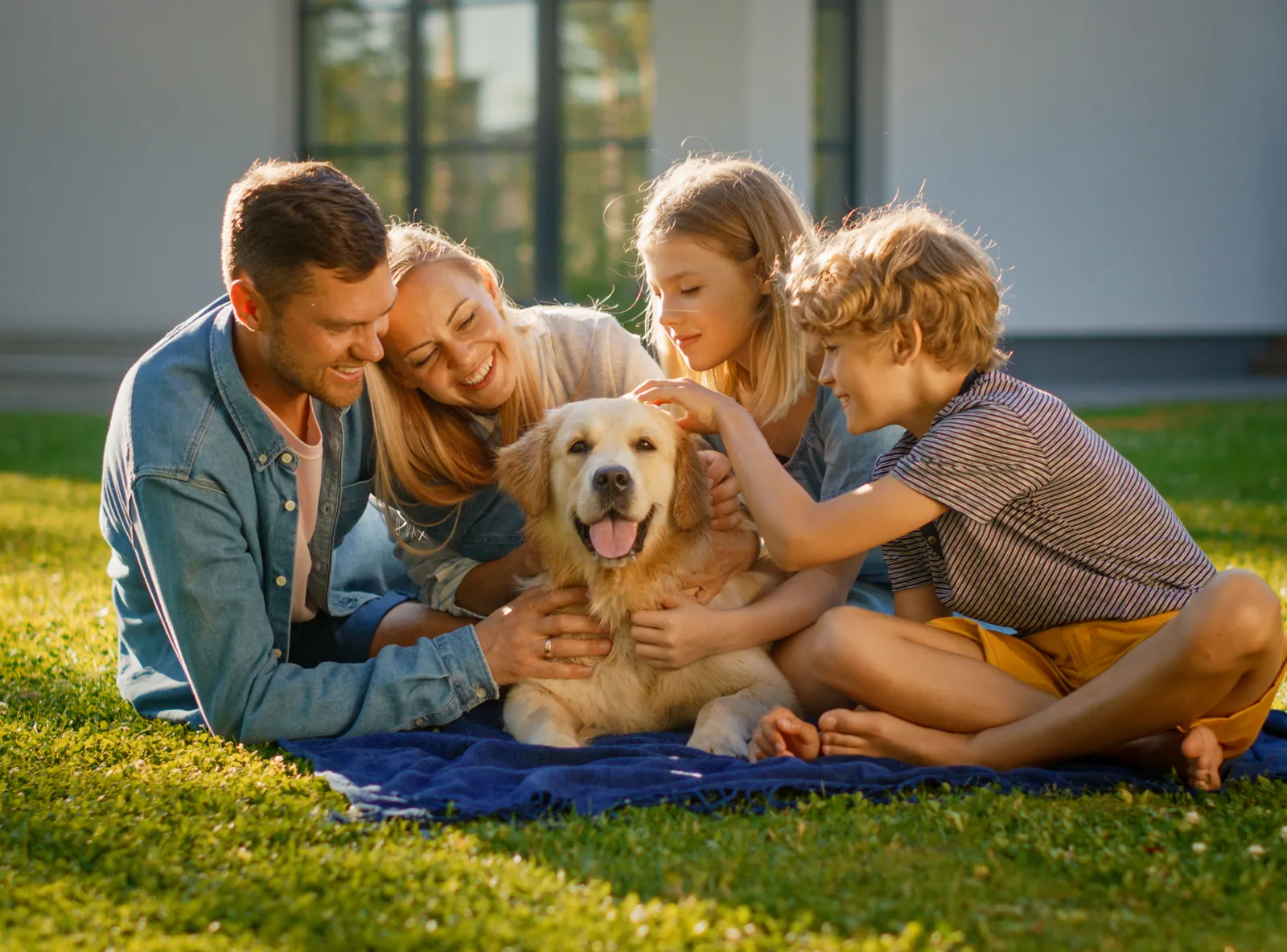 A family sits on a blanket in the sun, petting their golden retriever