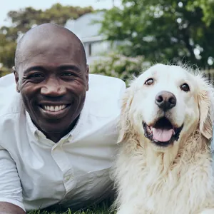 A smiling man lies on the grass beside his happy golden retriever.