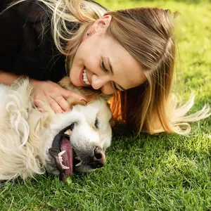 A woman lies cheek to cheek with her golden retriever on the grass, both grinning.
