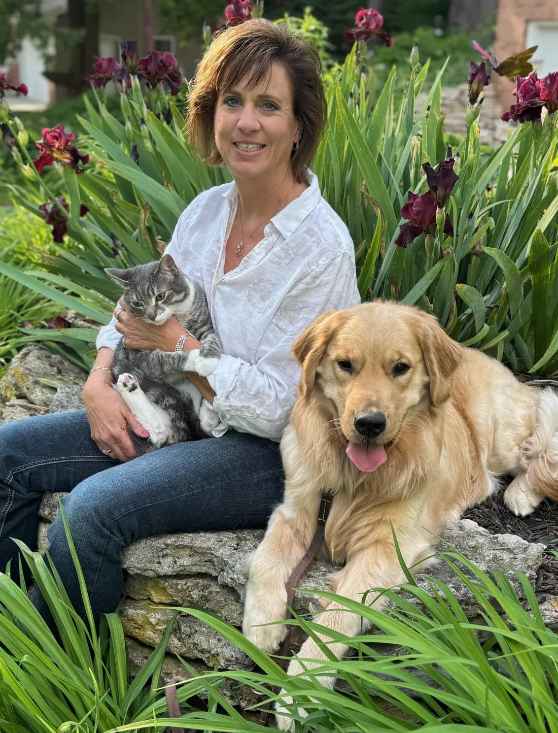 Dr. Karen Strickfaden sits in a garden with a gray cat on her lap and a golden retriever beside her